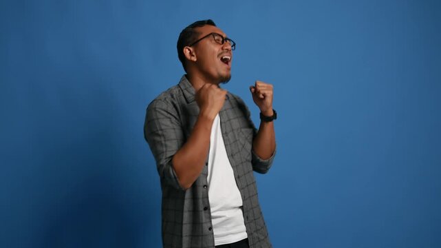 Happy asian adult man dancing celebrating success while raising fists isolated on blue background