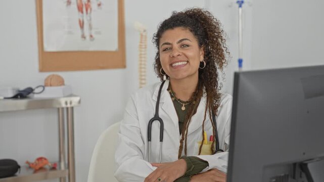 Woman doctor with stethoscope smiling, hand resting on desk and facing computer in building; compassionate care.