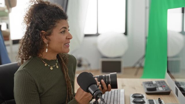 Woman holding lens speaks into microphone in studio while gesturing toward camera and gear; creative tutorial demo.