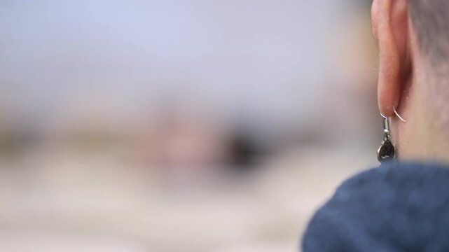Close-up of person's ear wearing a dangling earring. Person views a blurred scene ahead. Focus on detail of ear and clothing texture. Could be used for listening, concentration