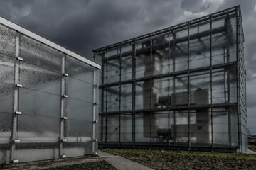 Nowoczesna architektura z przeszklonymi elewacjami w deszczowej scenerii. Minimalistyczne budynki o industrialnym charakterze na tle ciemnych, burzowych chmur, podkreślające współczesny design i surow © Lena