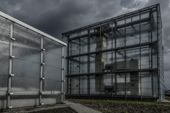 Nowoczesna architektura z przeszklonymi elewacjami w deszczowej scenerii. Minimalistyczne budynki o industrialnym charakterze na tle ciemnych, burzowych chmur, podkreślające wsp&oacute;łczesny design i surow