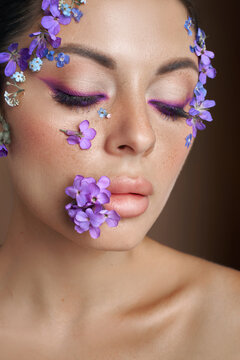 A model stands in a studio with purple flowers on her face and around her eyes. The setting has soft lighting that highlights her features. Her expression shows focus while getting ready