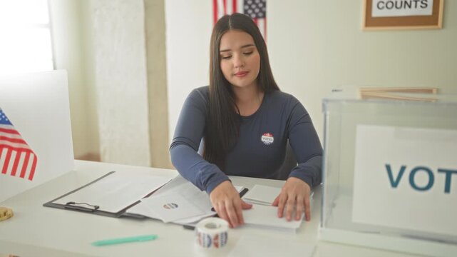 Woman placing ballot into clear ballot box with hand at voting table in building, wearing an i voted sticker and us flag visible on wall; civic duty pride.