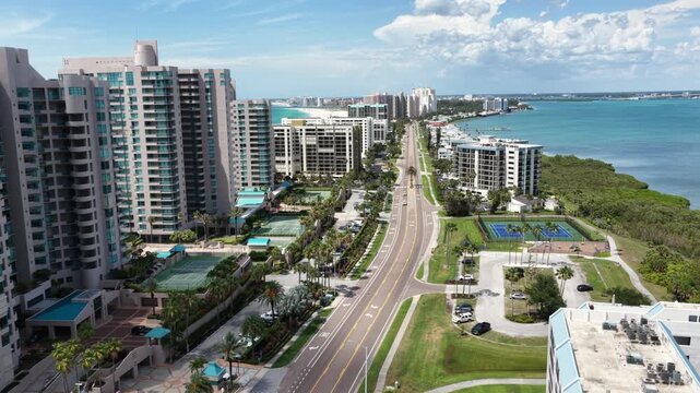 Palm-lined avenue runs between high-rise condo towers, tennis courts and parking lots bordering calm intracoastal waters under summer sky. Clearwater Beach, Florida, USA. Aerial drone rise wide shot