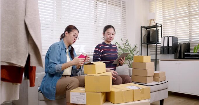 Asian women small business partners preparing online shopping parcel box, sealing packages, checking order details, arranging shipping boxes, managing e-commerce fulfillment process inside home