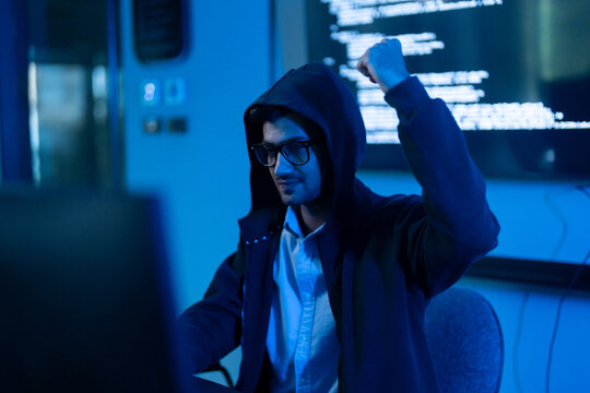 Successful male hacker in hoodie raising fist celebrating victory in dark blue room with computer code screen, Cybercrime attack, cybersecurity triumph, data breach and digital privacy breach