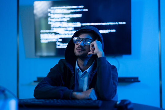 Thoughtful male hacker in hoodie and glasses sitting in dark blue room with computer code on screen, Cybercrime, cybersecurity threat, dark web, data breach, malicious software and digital privacy