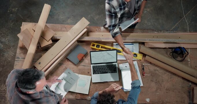 Carpenter, team and handshake in factory with paperwork, design update and agreement. Above, men and shaking hands at laptop for woodworking project, feedback and task delegation for productivity