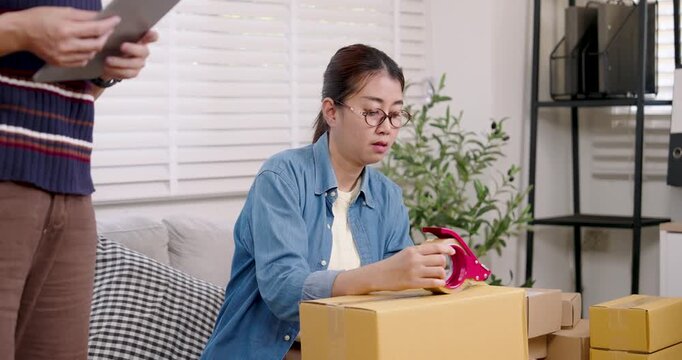 Asian female entrepreneur sealing parcel box with tape, finalizing packaging step, preparing online shopping parcel box for shipping inside home based e-commerce business workspace