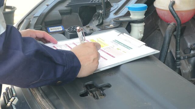 A mechanic holding a pen and writing on a vehicle inspection form checklist on a clipboard, with an open car engine compartment in the background. Close-up with selective focus.