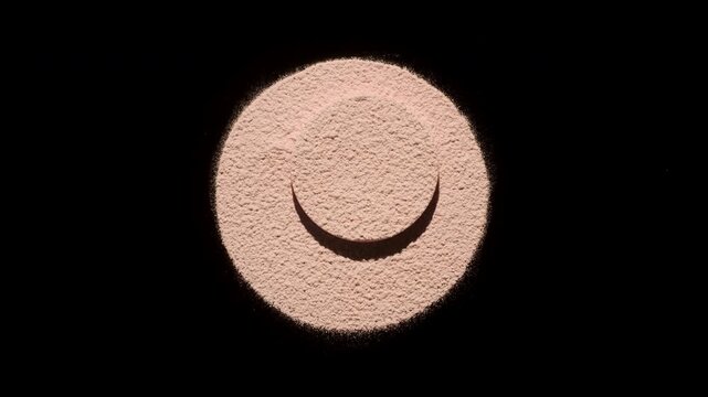 Top-down shot of beige sifted powder in a circular shape with a smaller round platform at the center intercepting moving light that casts shadows against black background | Concept for matte powder