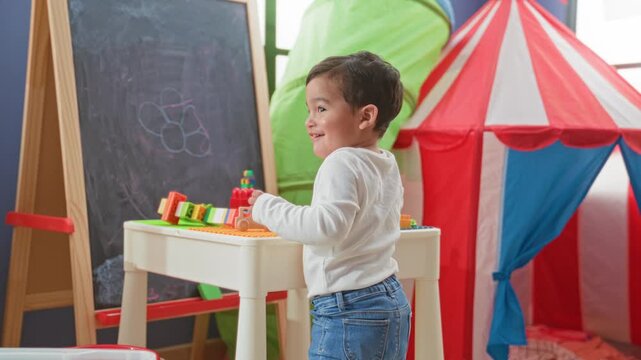 Boy at play table stacking toy blocks, hand reaching toward colorful bricks beside a chalkboard and play tent in building; curiosity and play.