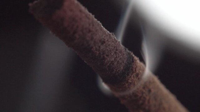 Macro view of smoldering incense stick releasing wisps of smoke, highlighting burnt ash texture, combustion process, and aromatic detail against blurred background