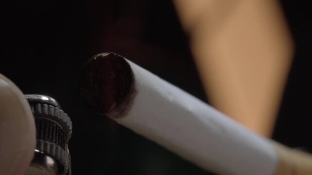 Macro view of lit cigar and metal lighter during ignition, highlighting charred tobacco, burning tip, combustion process, and warm illumination in close detail.