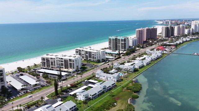 High-rise condos line Clearwater Beach island, turquoise Gulf surf meets white sand, intracoastal waterway and mangroves edge inland shore. Clearwater Beach, Florida, USA. Aerial drone flyover shot