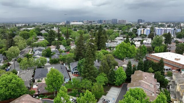 Storm clouds gather above red-tile townhome rooftops and trees, dense residential community giving way to San Jose skyline on distant horizon. San Jose, California, USA. Aerial drone flyover shot