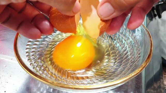 A person cracking a fresh egg into a clear glass bowl with a gold rim on a kitchen counter