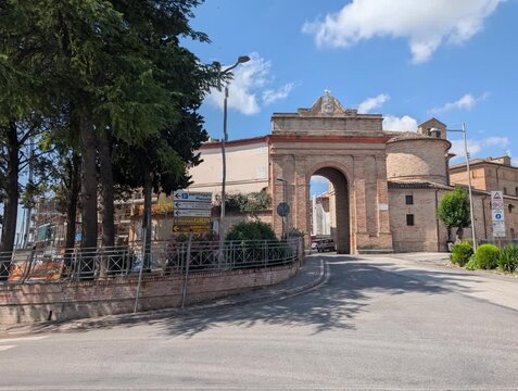 L&rsquo;ingresso storico di Urbisaglia si apre con l&rsquo;arco in mattoni e la strada che va verso il centro. Una scena luminosa che unisce architettura antica e vita quotidiana marchigiana.