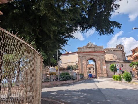 L&rsquo;ingresso storico di Urbisaglia si apre con l&rsquo;arco in mattoni e la strada che va verso il centro. Una scena luminosa che unisce architettura antica e vita quotidiana marchigiana.