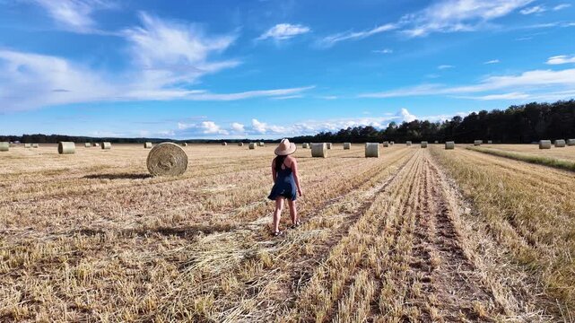 Woman in straw hat wanders through open stubble field, hay bale beside her, Maine