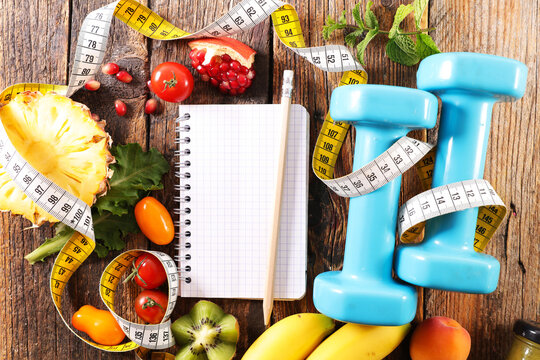 Fitness dumbbells with measuring tape and healthy food around blank notebook on wooden background. Diet food concept