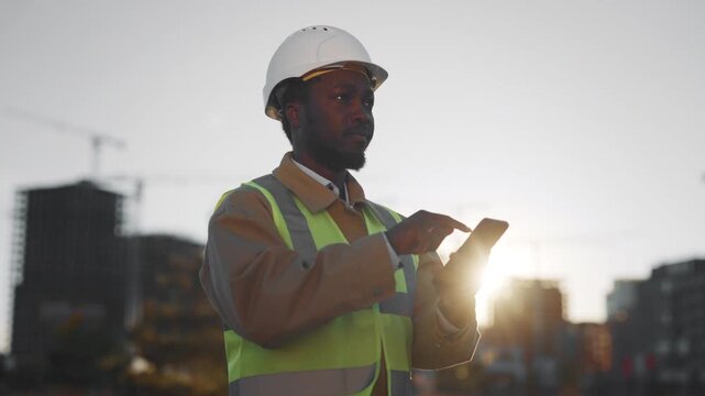 African builder chatting by mobile app in smartphone in construction site. Portrait of foreign professional foreman or engineer standing before building under construction, african american ethnicity