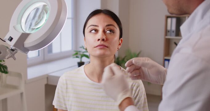 Woman client coming to consultation to plastic surgeon or aesthetic dermatologist, discussing correction of face, sitting under lamp, listening recommendations. Plastic surgery and cosmetology
