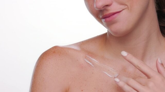 Big closeup of fair-skinned freckled Spanish woman applies moisturizing cream on her clavicle on white bg | rejuvenating cream commercial