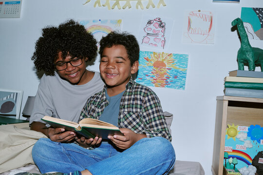 Black teenager and Black child reading book together, smiling and bonding at home beneath childrens artwork. Image supporting family literacy, homeschooling, sibling care marketing