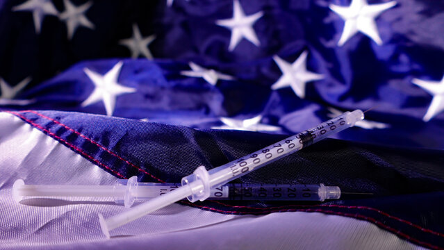 Syringes lay on a folded American national flag representing discussions and concerns surrounding US healthcare, medical treatment, and public health policy