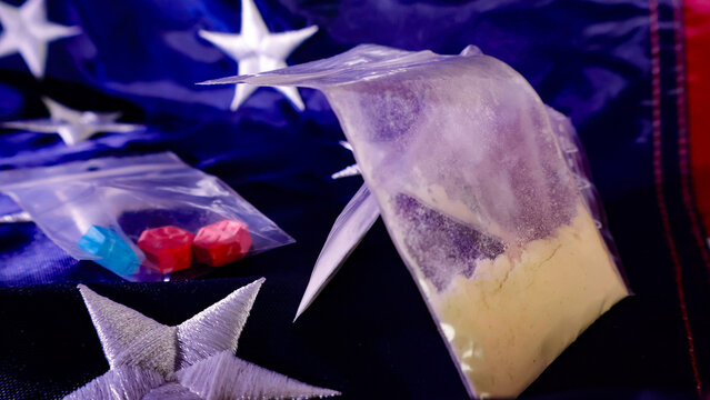 Illicit drugs and colorful pills packaged in clear plastic bags are spread across a background resembling a United States flag, symbolizing issues of drug trafficking and addiction