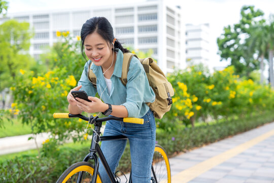 Asian woman stands near a bicycle in a city park and is using her cell phone.