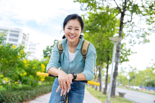 Asian woman in casual clothes, carrying a backpack, smiles warmly while cycling in the park.