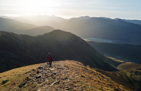 A female hiker and their dog walking down from the summit of Grasmoor with Whiteless Pike and Robinson in the distance on a bright winters morning in the Lake District, UK
