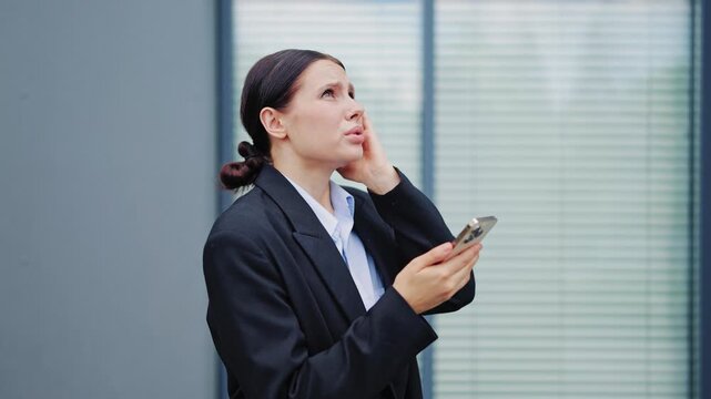 Worried businesswoman with a headache looks at her smartphone, reflecting confusion and stress. Appears to be processing challenging news or technical issues.
