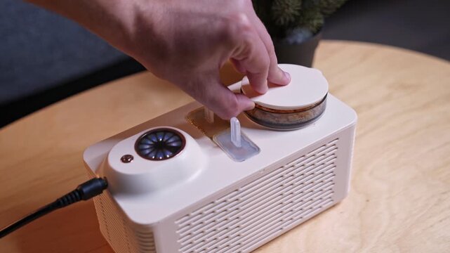 a hand removing the clear lid from a crumb tray on a white mini ice pop maker. The modern electric appliance sits on a wooden table inside a cozy home living room setting.