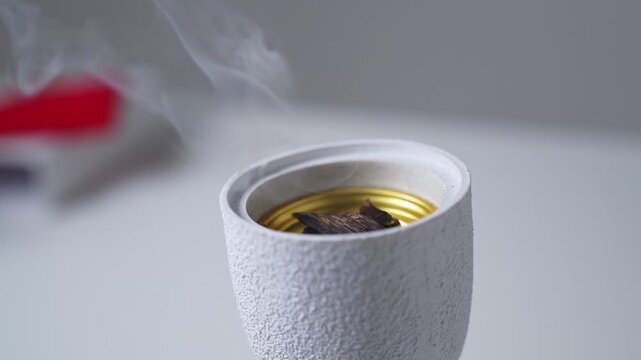 traditional oud wood burning inside a modern white ceramic incense burner. Wisps of aromatic smoke rise from the gold interior against a bright indoor home setting