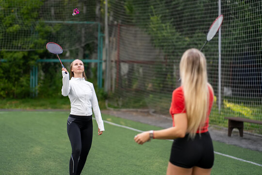 Two young women playing badminton on an outdoor green sports court. One holding a racket and shuttlecock, ready for a rally.