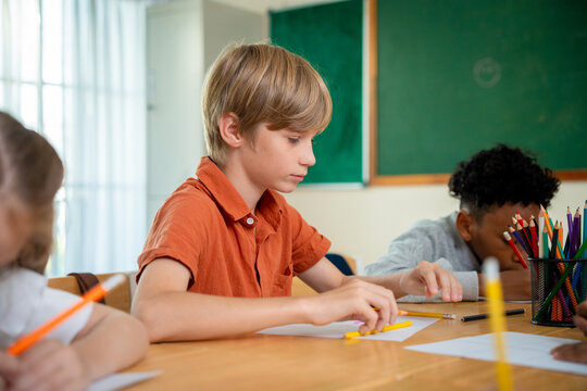 Primary school boy using a yellow pencil at his wooden desk during a classroom lesson. Schoolchild concentrating on drawing or writing with colored pencils in a diverse elementary classroom.