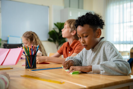 African American schoolboy concentrating on folding green origami paper at his classroom desk. Creative multiethnic student focusing on paper folding craft during an art and craft lesson.