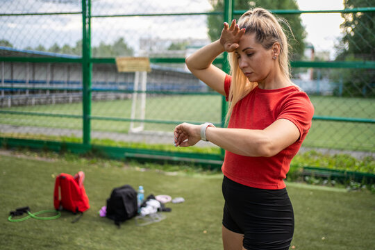 Tired young woman wiping sweat from forehead after workout and checking smartwatch. Post-exercise recovery and performance tracking.