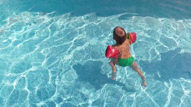 A kid in oversleeves learns to swim in a children's pool with clean water, funny moving his legs on a summer day