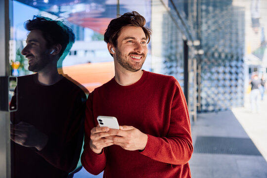 Smiling man using smartphone with wireless earbuds in the city