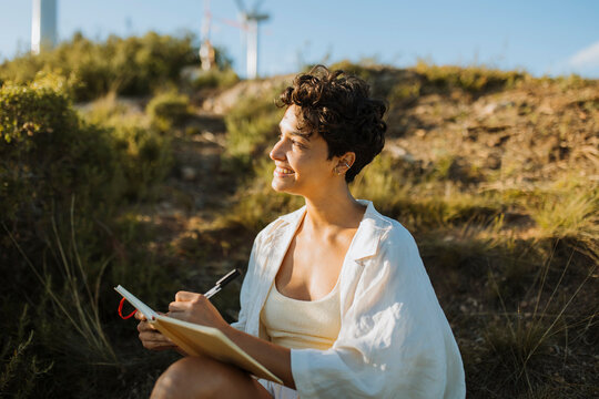Smiling woman holding pen and notebook while sitting on mountain