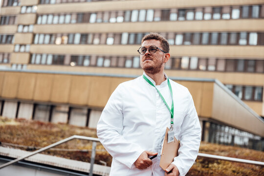Doctor standing outdoors by hospital in lab coat with clipboard