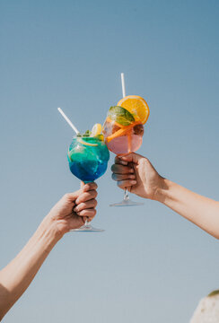 Two hands clink glasses of colourful cocktails against a blue sky