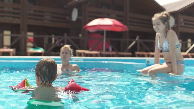 Boy in oversleeves swims in a pool of clear water with mom and sister on summer vacation