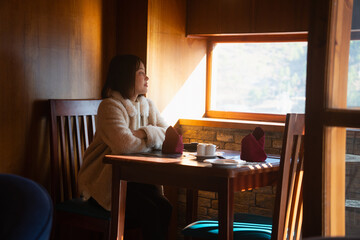 A young Asian woman in a warm cream fleece coat sitting at a dining table by the window, crossing her arms and looking out at the view, relaxing inside a cozy wooden restaurant or resort in winter © Oil And Den