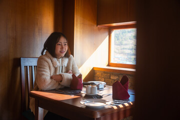 A young Asian woman wearing a warm cream fleece coat sitting at a dining table with arms crossed, smiling and relaxing by a window with warm sunlight inside a cozy wooden restaurant © Oil And Den